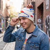 A man wearing a cream knit beanie with colorful cloud and star patterns, a blue denim jacket, and a graphic t-shirt, standing in an outdoor urban alleyway.