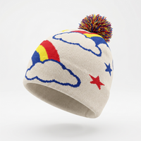 A knit beanie hat in warm off-white with a multicolor rainbow cloud pattern, blue stars, and a matching multicolor pom-pom on top, shown against a light gray studio background.