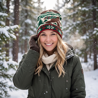 A smiling blonde woman in a snowy forest wearing a festive knit beanie with a green, brown, and red holiday pattern, a cream knit scarf, and an olive green winter jacket.