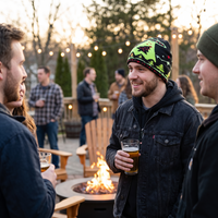 Three men gathered outdoors around a fire pit at dusk, with the central person wearing a dark denim jacket and a bright green and black knit beanie while holding a drink.