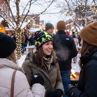 A smiling woman in a winter outdoor setting wears a black knit beanie with green and yellow tree patterns, a brown puffer jacket, and a beige scarf. Other people in winter clothing are visible around her, with snow, trees, and warm string lights in the background.