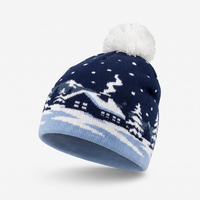 A navy blue knit beanie with a white pom-pom on top, featuring a knitted winter village scene with snow-covered trees and houses. The cuff of the beanie is light blue.