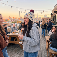 A woman on a rooftop bar deck at sunset wearing a light gray hoodie and a cream beanie with colorful cartoon graphics and a red pom-pom. She is holding a wine glass and smiling, with other people, tables, and a city skyline in the background.