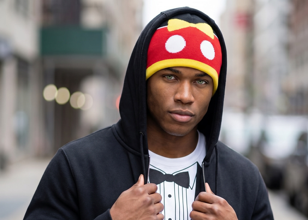 Person wearing a black hoodie with a mickey mouse beanie standing on a city street.